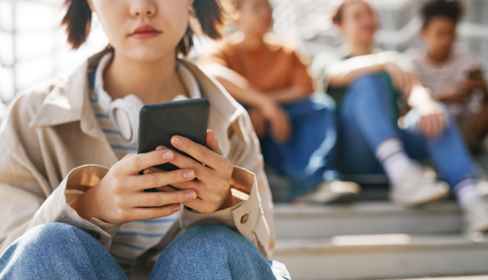 Young woman with headphones around her neck looking at her cell phone screen while sitting in the bleachers with a row of young people sitting in the background