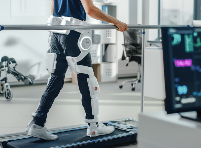 Person walking on treadmill with white electronic leg braces attaching a waist, with monitoring screen in the foreground showing vital signs