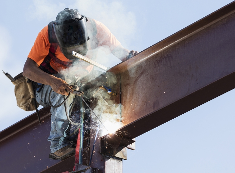 A worker is elevated above a building welding an I-beam. Exponent materials engineers consult on welding and fabrication. 
