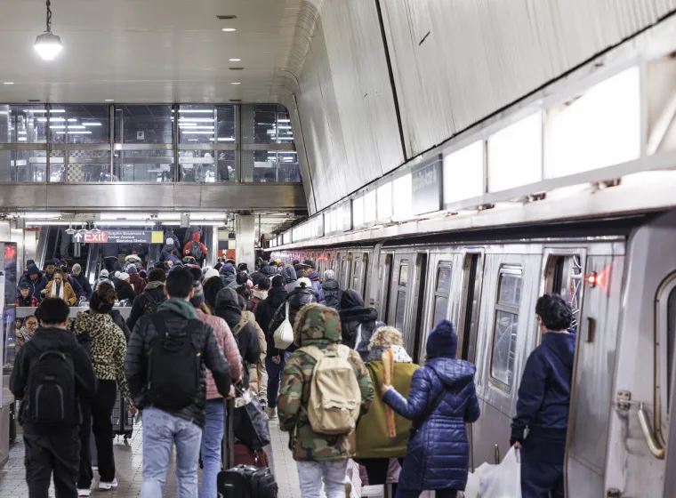 A large crowd of people waits on the platform and boards a subway train at Jamaica Station in Queens, New York City. The diverse group of commuters is dressed in winter clothing for the December weather. This scene captures the daily evening rush hour on the city's public mass transit system.