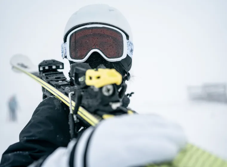 A close-up action shot of a skier wearing a white helmet and red-tinted goggles, gripping a yellow ski pole with a snowy, blurred background that conveys cold, movement, and outdoor sport.