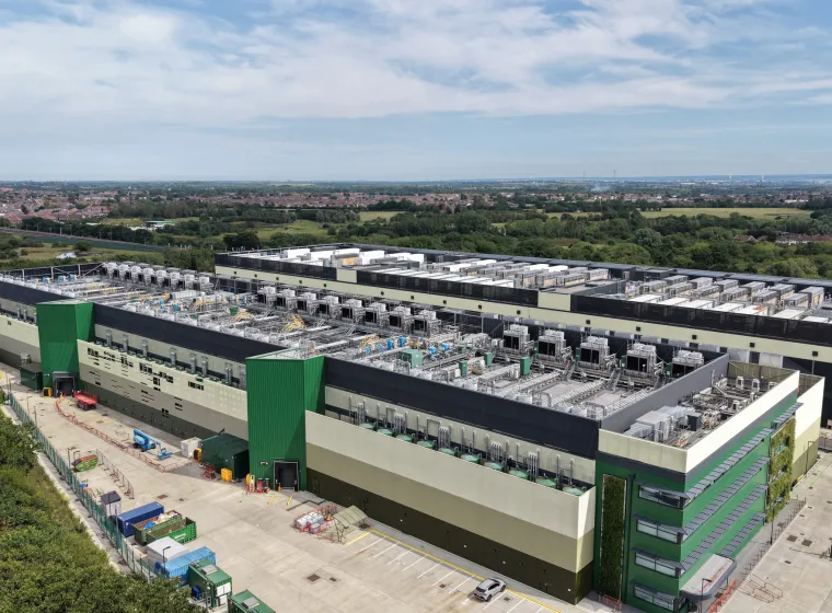 Aerial view shows a large, modern data center facility featuring green and beige facades. Extensive rooftop cooling units are prominent.