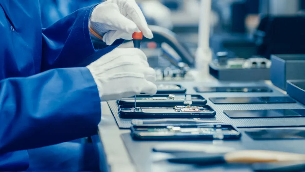 Close Up of a Female Electronics Factory Worker in Blue Work Coat and Protective Glasses Assembling Smartphones with Screwdriver. High Tech Factory Facility with more Employees in the Background.