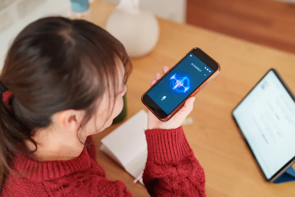 Young woman with cold and flu symptoms using a smartphone voice assistant to get health advice at home. She speaks into the phone while resting at a table with medicine, tissues, and a tablet nearby. Concept of telemedicine, digital healthcare, remote support, voice-controlled technology, home recovery, and modern health tools.