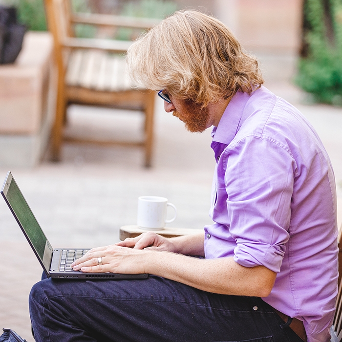 A person sitting outside, working on a laptop with cup of coffee. Exponent's engineering research advances next gen computer electronics.