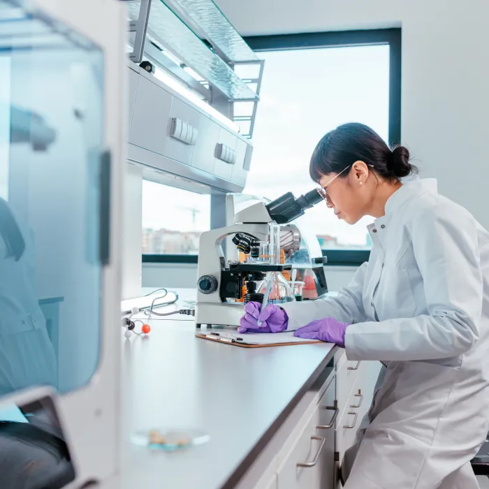 A female scientist is sitting in her lab and looking trough the microscope, she is writing down notes about her discoveries after looking at samples