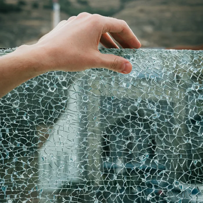 A human hand touches the web-like pattern of shattered safety glass, with blurred natural landscape in the background. The image captures a moment of stillness and curiosity.