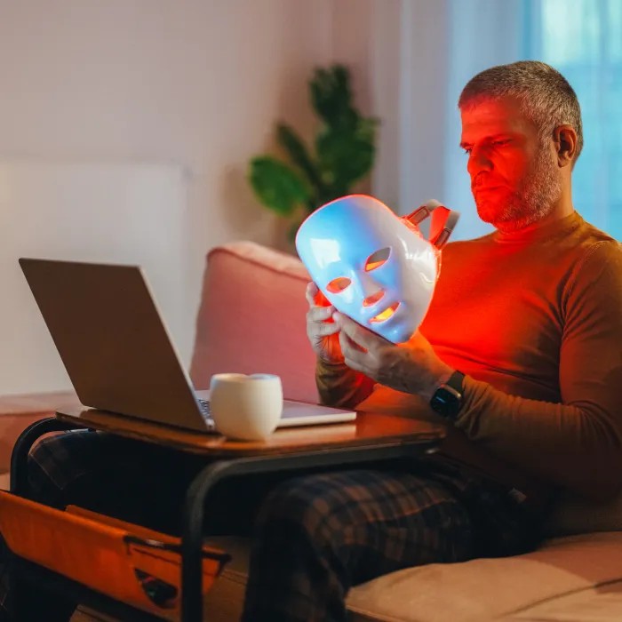 A middle-aged man sits comfortably on the sofa, wearing a red light therapy mask while working on his laptop and enjoying a coffee at home. The scene highlights modern wellness trends, remote work, and the integration of health technology in daily life.