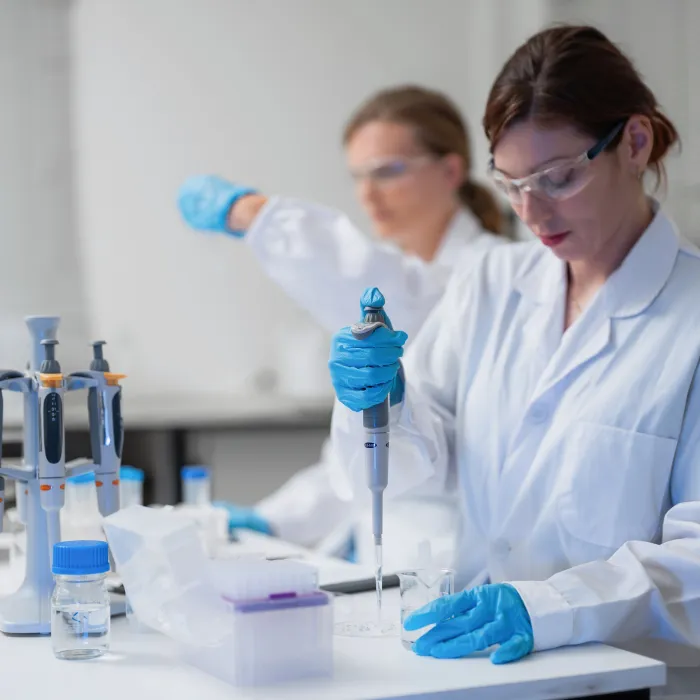  Scientist in white lab coat uses pipette with blue gloves. Another researcher gestures with a pipette in the background. Lab table holds multiple pipettes, containers, and a water bottle