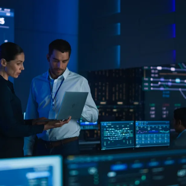 Project Leader and Chief Executive Discuss Data Shown on a Laptop. In Background Big Screen Showing Infographics of Infrastructure, Charts, System Analysis. Telecommunications Control Room