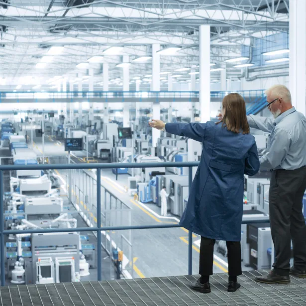 Experienced Male and Female Engineers Standing on a Platform with Their Back to Camera, Using Laptop Computer and Discussing Production at a Modern AI Automated Electronics Manufacture