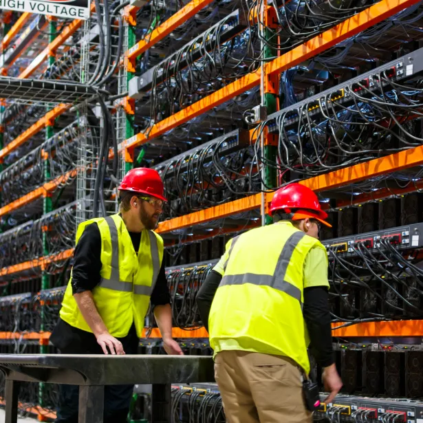 Two technicians wearing PPE walking past racks of equipment in a data center for cryptocurrency mining, cloud services and AI computing in a large, temperature controlled warehouse in a remote location in Stutsman County, North Dakota. One of them is pushing a cart.