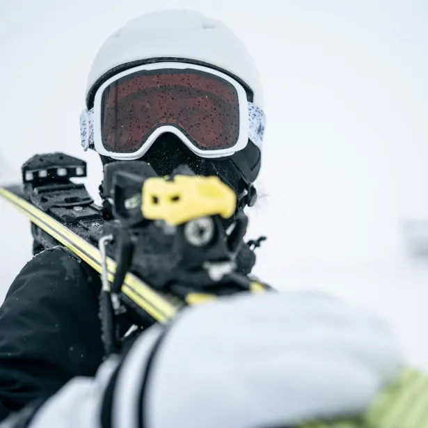 A close-up action shot of a skier wearing a white helmet and red-tinted goggles, gripping a yellow ski pole with a snowy, blurred background that conveys cold, movement, and outdoor sport.