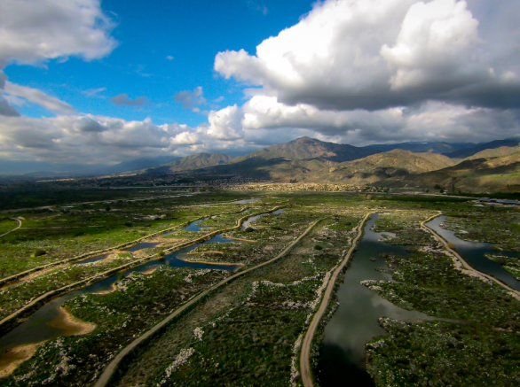 Aerial view of green valley with ponds/marshes and two-lane roads running through against backdrop of mountains and blue, cloud-covered sky