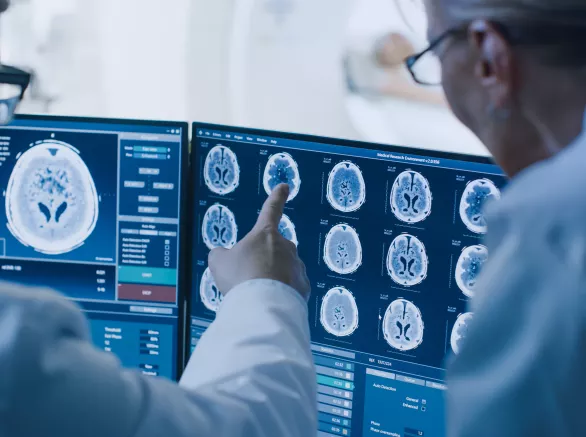 In Control Room Doctor and Radiologist Discuss Diagnosis while Watching Procedure and Monitors Showing Brain Scans Results, In the Background Patient Undergoes MRI or CT Scan Procedure