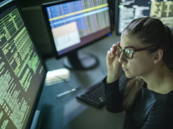 A young woman is seated at a desk surrounded by monitors displaying data, she is contemplating in this dark, moody office.