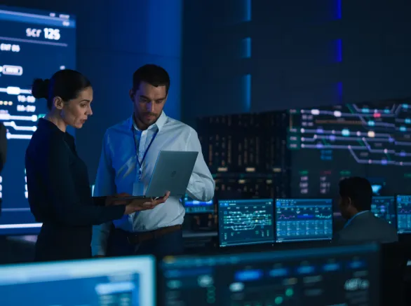 Project Leader and Chief Executive Discuss Data Shown on a Laptop. In Background Big Screen Showing Infographics of Infrastructure, Charts, System Analysis. Telecommunications Control Room