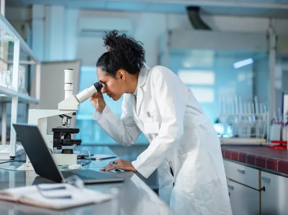  Female Scientist Looking Under Microscope And Using Laptop In A Laboratory