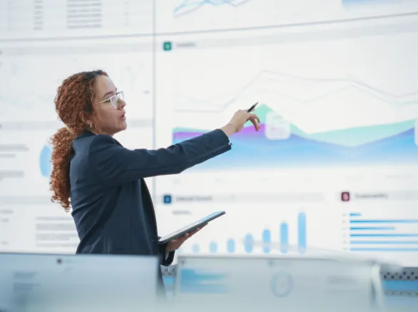 Hispanic Female Senior Data Scientist Reviewing Reports Of Risk Management Department On Big Digital Screen In Monitoring Room. Diverse Consulting Company Employees Working Behind Desktop Computers.