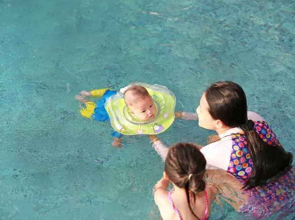  Mother and daughter training for infant baby boy in swimming suit floating in pool with safety by baby neck floats. 