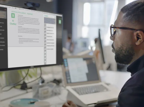 Stock image showing a black man’s face looking into a computer screen in an open plan working office. Type is being added to the screen by an Artificial intelligence, AI, chatbot.