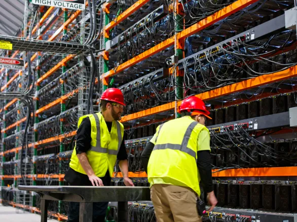 Two technicians wearing PPE walking past racks of equipment in a data center for cryptocurrency mining, cloud services and AI computing in a large, temperature controlled warehouse in a remote location in Stutsman County, North Dakota. One of them is pushing a cart.