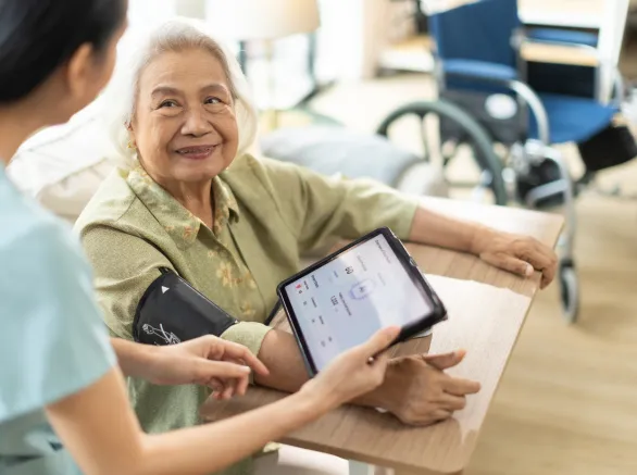  Woman asian nurse taking blood pressure of a senior woman patient at home.Home care healthcare professional using digital tablet 