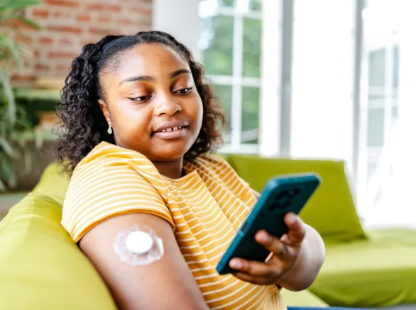 A young black woman sitting on a green couch checks her phone while managing her type 1 diabetes. A sensor is attached to her arm, indicating her health monitoring approach at home.