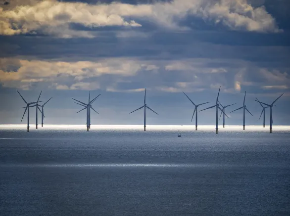 Offshore wind farm set against a dramatic sky with dense clouds. A narrow strip of sunlight illuminates the ocean surface in front of the wind turbines, creating a striking contrast. The turbines are evenly spaced, with large blades harnessing wind energy. The deep blue sea is calm, and the atmosphere suggests a peaceful, yet dynamic environment. An isolated small boat appears near the turbines, highlighting the scale of the scene.