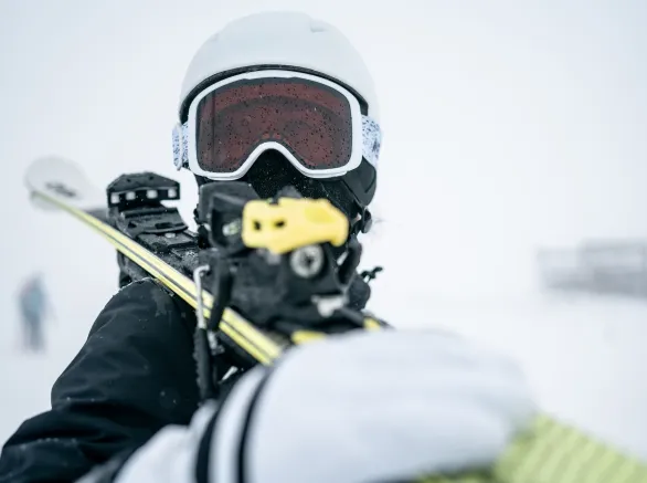 A close-up action shot of a skier wearing a white helmet and red-tinted goggles, gripping a yellow ski pole with a snowy, blurred background that conveys cold, movement, and outdoor sport.