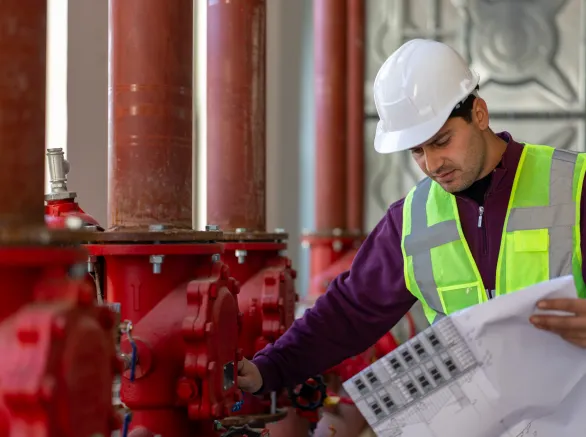 Engineer wearing a hard hat and safety vest, inspecting red industrial fire sprinkler pipes and checking blueprints, ensuring system integrity and safety in a commercial building