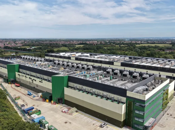 Aerial view shows a large, modern data center facility featuring green and beige facades. Extensive rooftop cooling units are prominent.