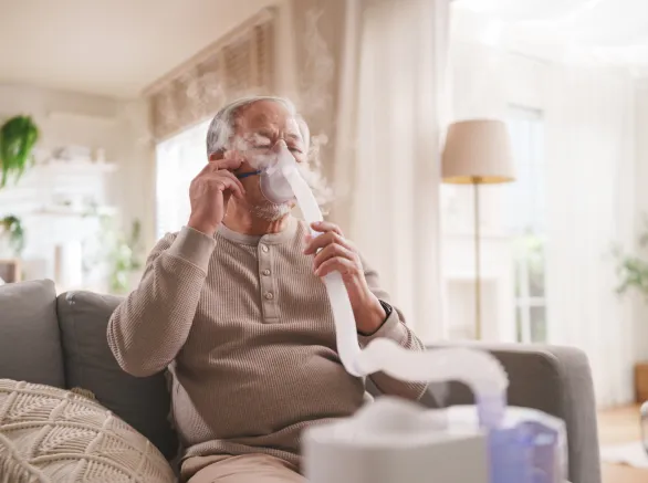 Senior Asian man uses a nebulizer mask for respiratory therapy while receiving inhalation treatment at home.