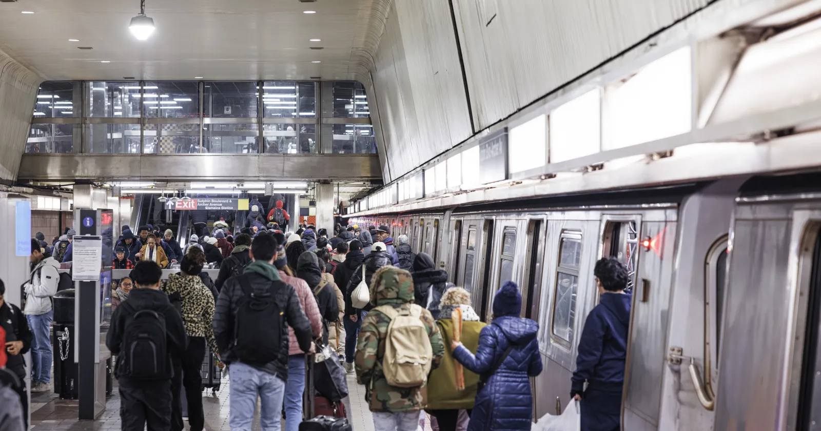A large crowd of people waits on the platform and boards a subway train at Jamaica Station in Queens, New York City. The diverse group of commuters is dressed in winter clothing for the December weather. This scene captures the daily evening rush hour on the city's public mass transit system.