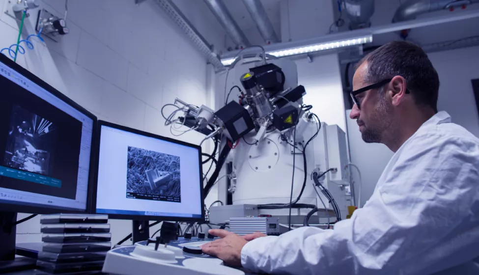 Sideview of man wearing glasses in a white lab coat sitting at a desk viewing objects on dual monitors with equipment in the background