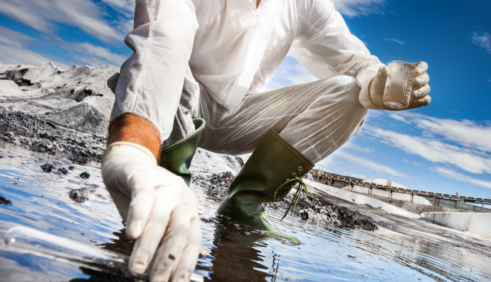 Field scientist crouched down in a shallow puddle holding a beaker and test tube, gathering a sample of the water.
