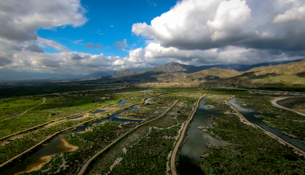 Aerial view of green valley with ponds/marshes and two-lane roads running through against backdrop of mountains and blue, cloud-covered sky