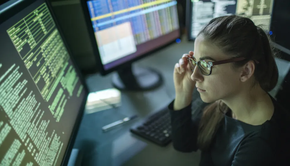 A young woman is seated at a desk surrounded by monitors displaying data, she is contemplating in this dark, moody office.