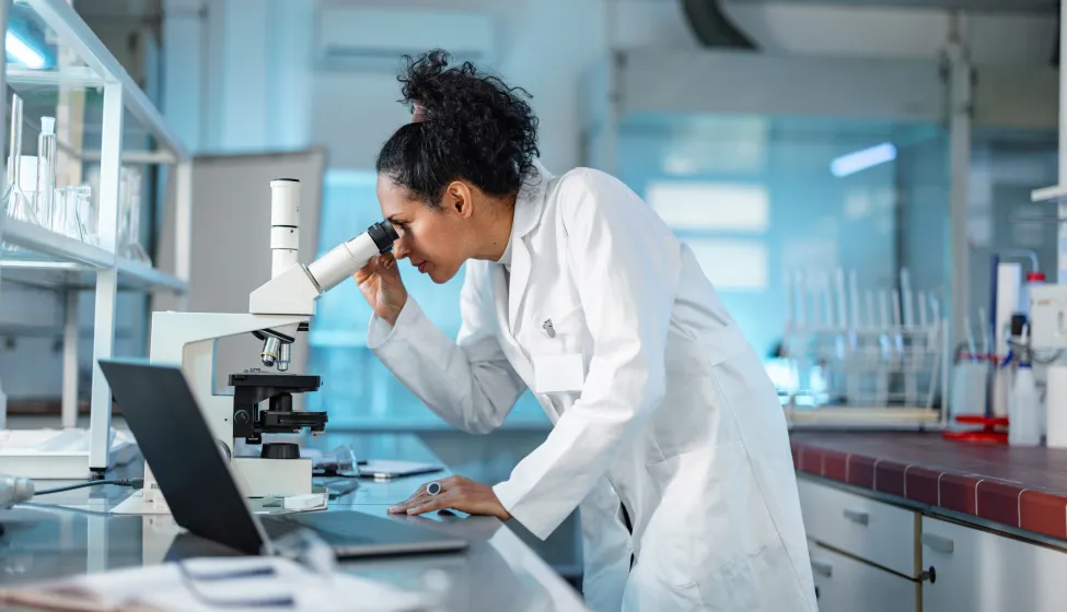  Female Scientist Looking Under Microscope And Using Laptop In A Laboratory