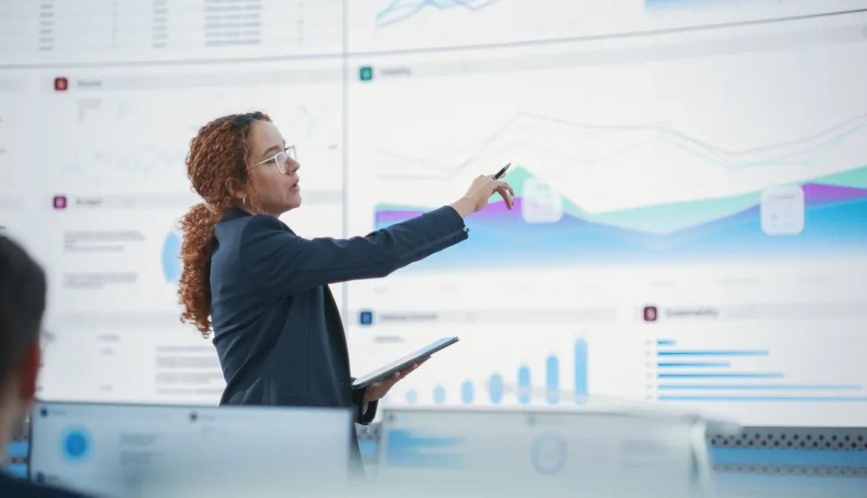 Hispanic Female Senior Data Scientist Reviewing Reports Of Risk Management Department On Big Digital Screen In Monitoring Room. Diverse Consulting Company Employees Working Behind Desktop Computers.