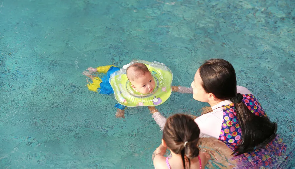  Mother and daughter training for infant baby boy in swimming suit floating in pool with safety by baby neck floats. 