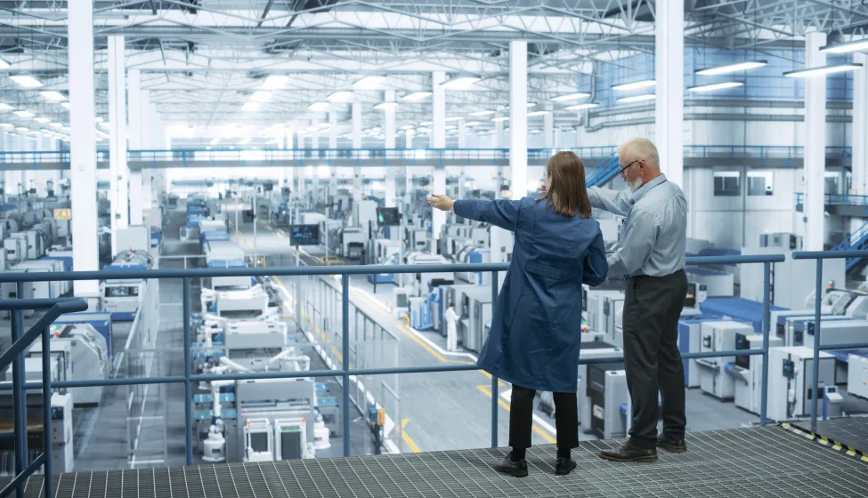 Experienced Male and Female Engineers Standing on a Platform with Their Back to Camera, Using Laptop Computer and Discussing Production at a Modern AI Automated Electronics Manufacture