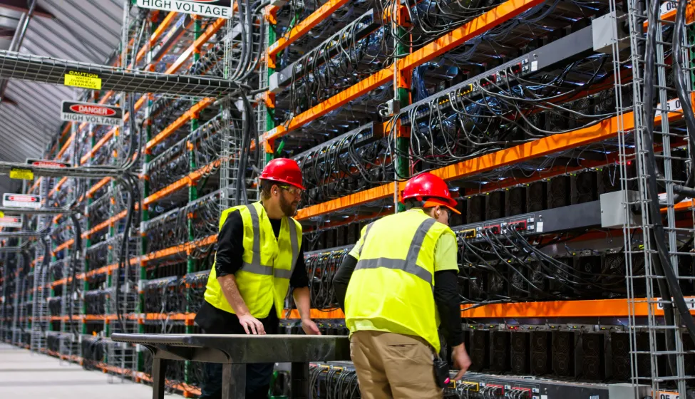 Two technicians wearing PPE walking past racks of equipment in a data center for cryptocurrency mining, cloud services and AI computing in a large, temperature controlled warehouse in a remote location in Stutsman County, North Dakota. One of them is pushing a cart.