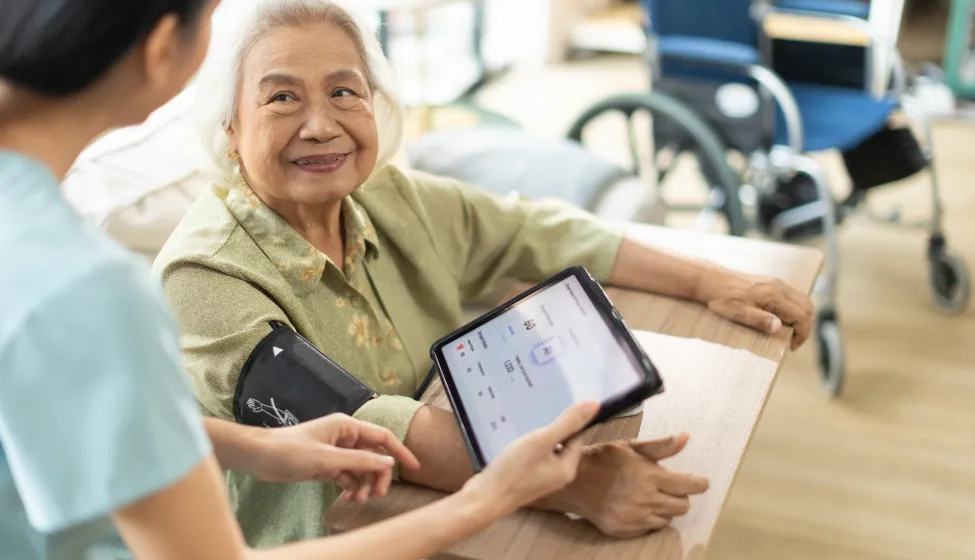  Woman asian nurse taking blood pressure of a senior woman patient at home.Home care healthcare professional using digital tablet 