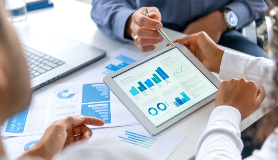 Close up of three people looking at financial data with graphs and charts. All their hands can be seen and one person is pointing with a pen. There is paperwork on the desk showing more finance information
