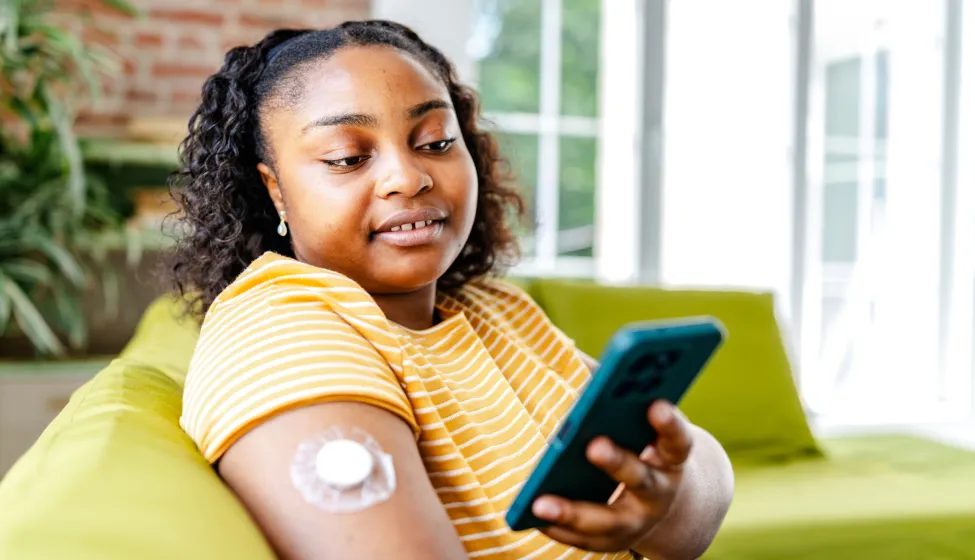 A young black woman sitting on a green couch checks her phone while managing her type 1 diabetes. A sensor is attached to her arm, indicating her health monitoring approach at home.