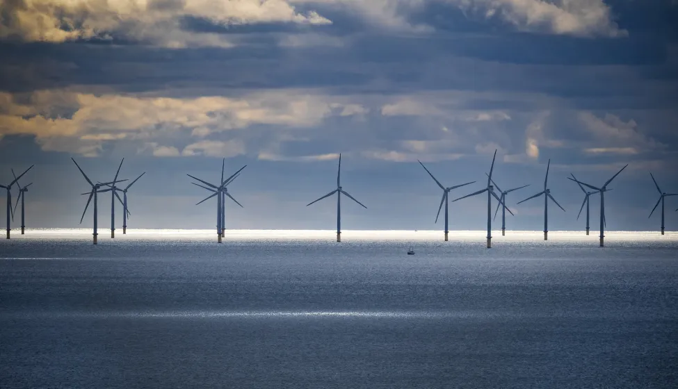 Offshore wind farm set against a dramatic sky with dense clouds. A narrow strip of sunlight illuminates the ocean surface in front of the wind turbines, creating a striking contrast. The turbines are evenly spaced, with large blades harnessing wind energy. The deep blue sea is calm, and the atmosphere suggests a peaceful, yet dynamic environment. An isolated small boat appears near the turbines, highlighting the scale of the scene.