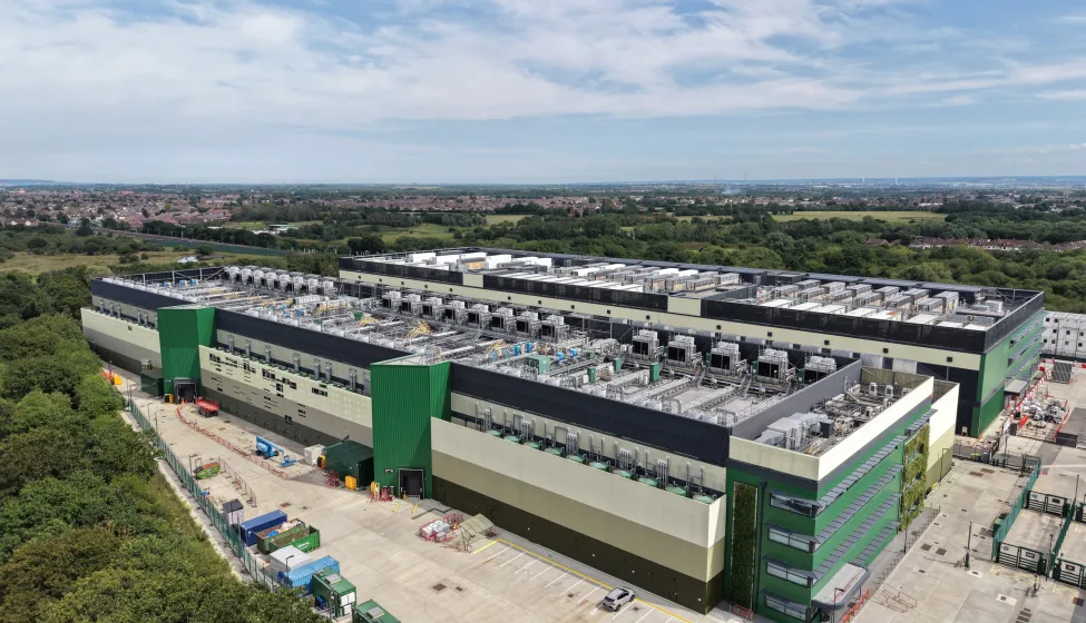 Aerial view shows a large, modern data center facility featuring green and beige facades. Extensive rooftop cooling units are prominent.