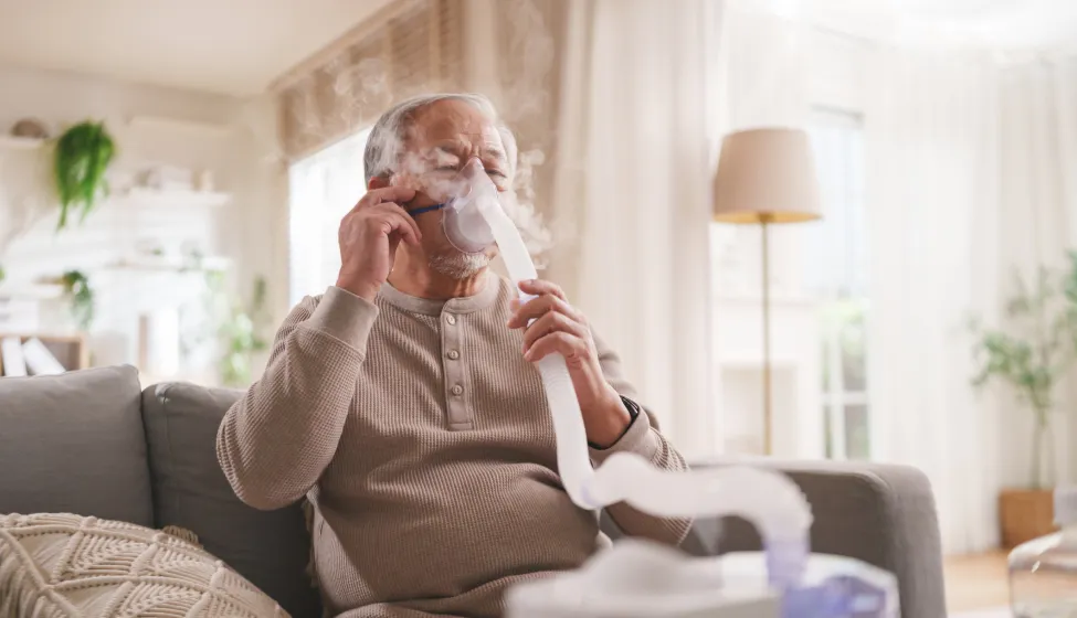 Senior Asian man uses a nebulizer mask for respiratory therapy while receiving inhalation treatment at home.