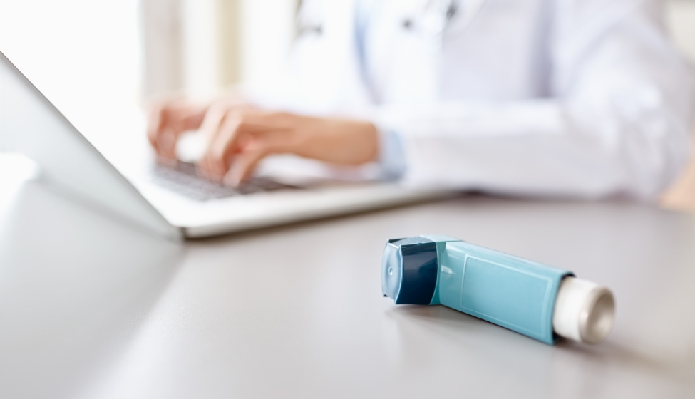 Inhaler on a desk with a medical professional using a laptop in the background